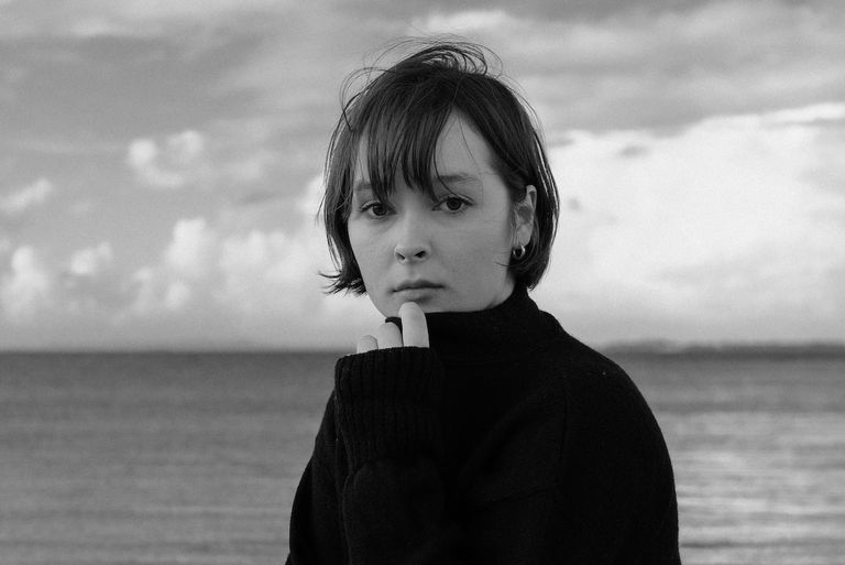 Black and white portrait of singer-songwriter Lottie McLeod in a high-neck sweater standing by the ocean under a cloudy sky.